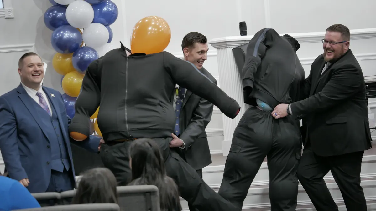 Bus workers in padded suits and balloon helmets racing in a game at the front of the auditorium