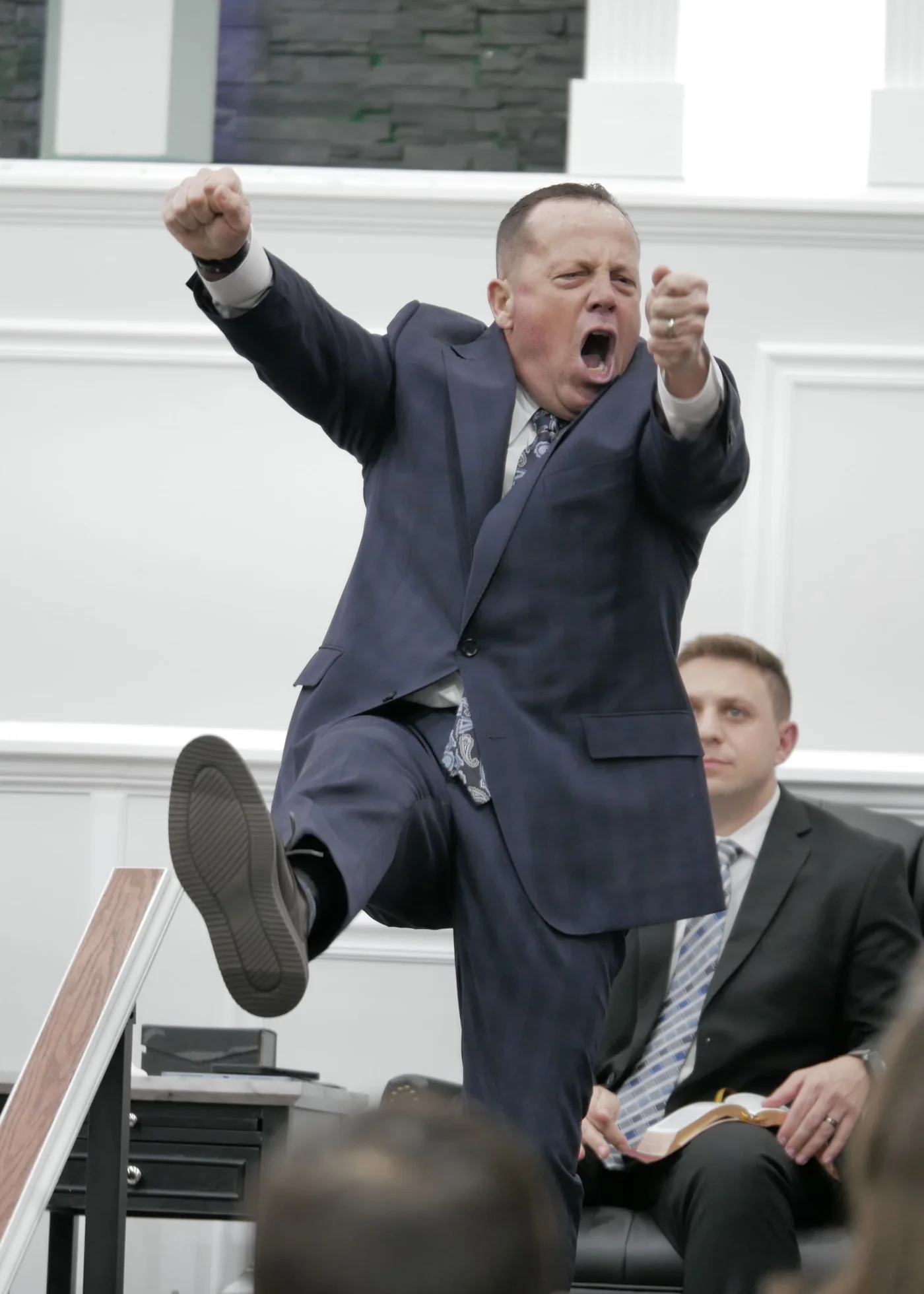 A preacher kicking his leg up and shouting with both fists out during a sermon