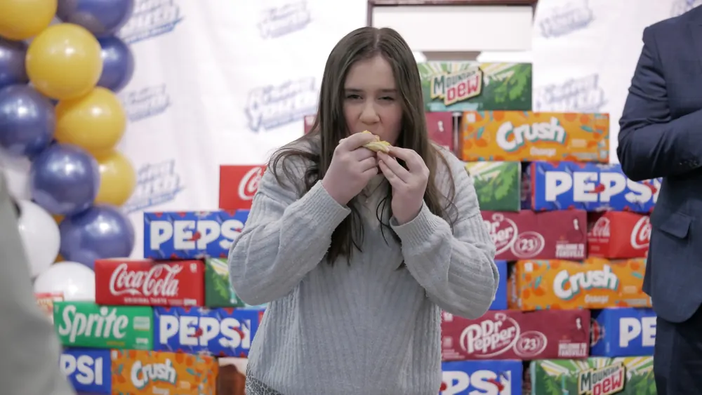 A teen taking part in an eating contest in front of a wall of soda boxes
